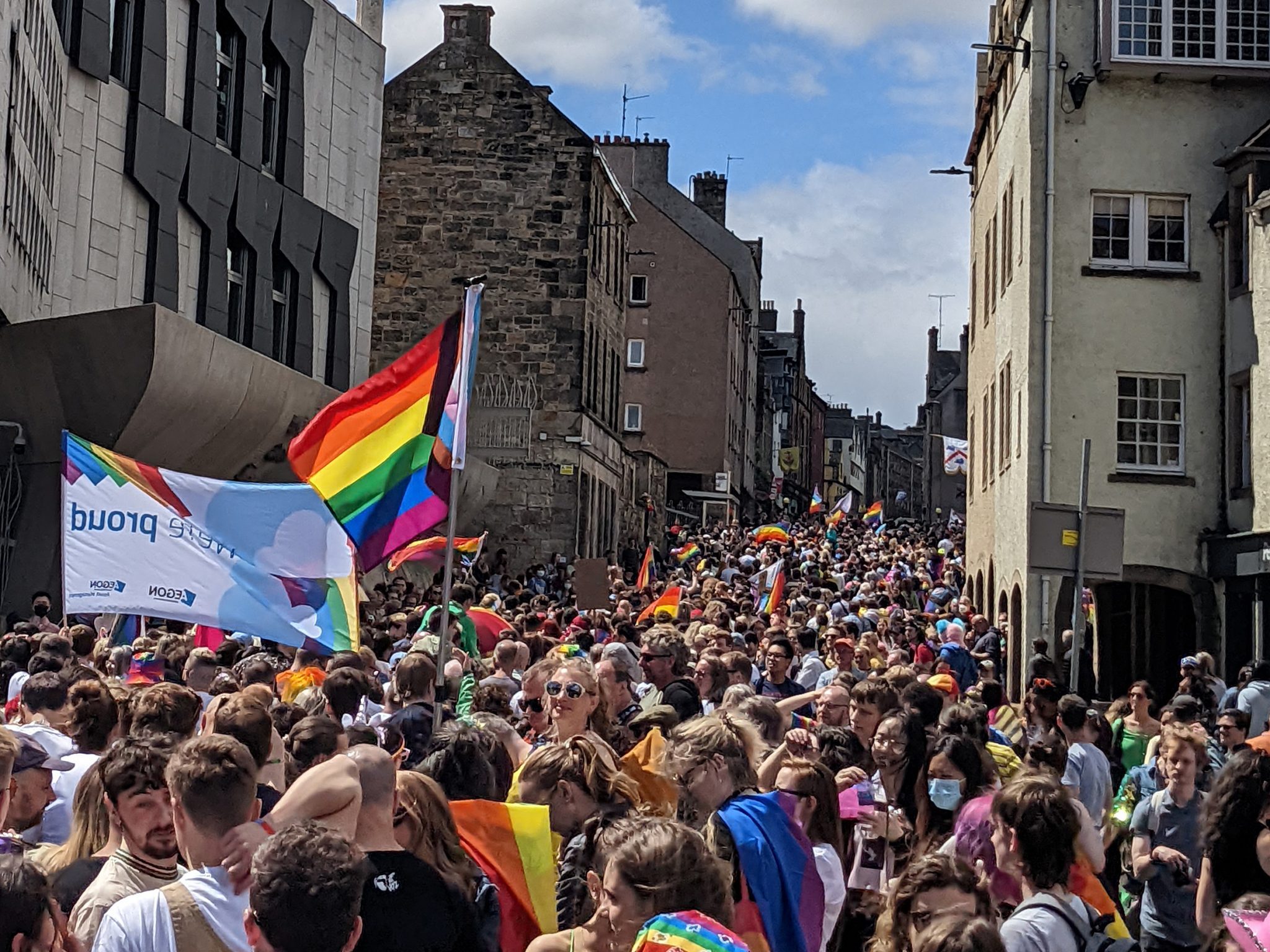 A large crowd of people going into the distance up a road up a hill with lots of pride and progress flags being waved.