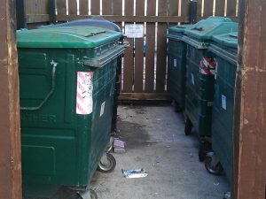 Large green recycling bins enclosed by brown wooden fencing.