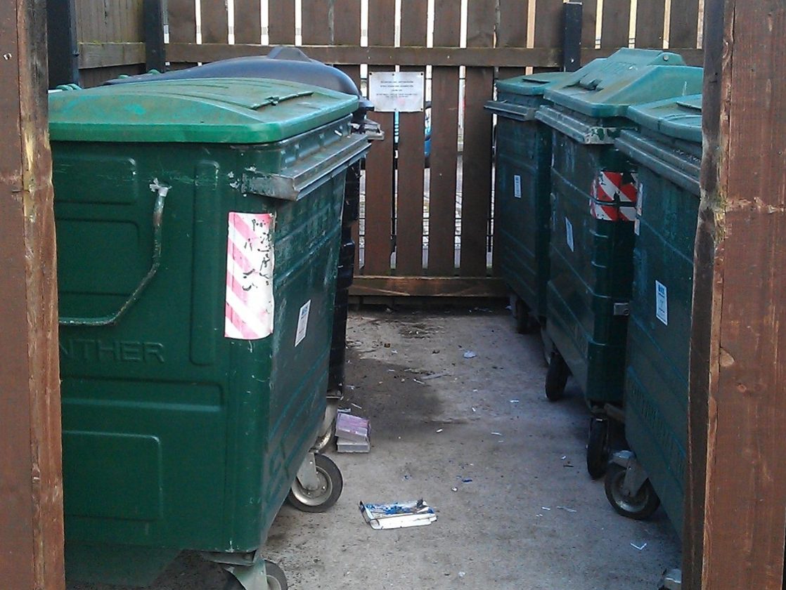 Large green recycling bins enclosed by brown wooden fencing.