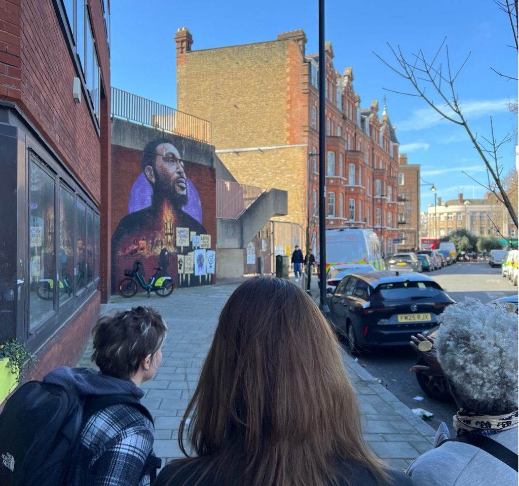 Three people look into the distance as they see a mural painted on the side of a building which depicts singer Marvin Gaye.