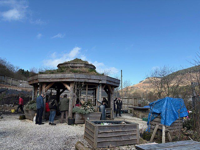 A large wooden pavilion sits on grey gravel, against a hillside backdrop with a bright blue sky.