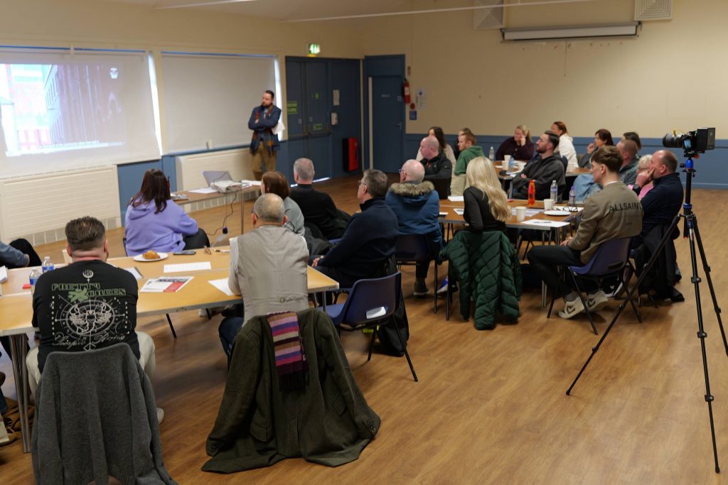 A crowd of people sat around tables watching a video during a consultation meeting.