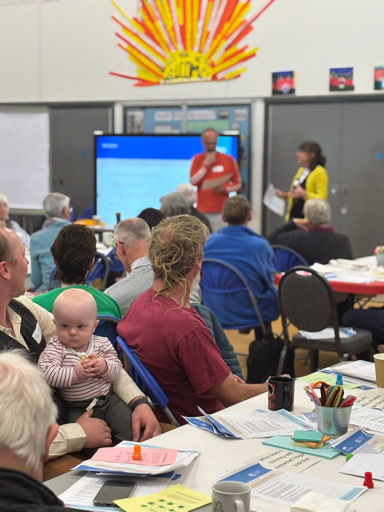 A crowd of people watching two adults give a presentation on a digital screen.